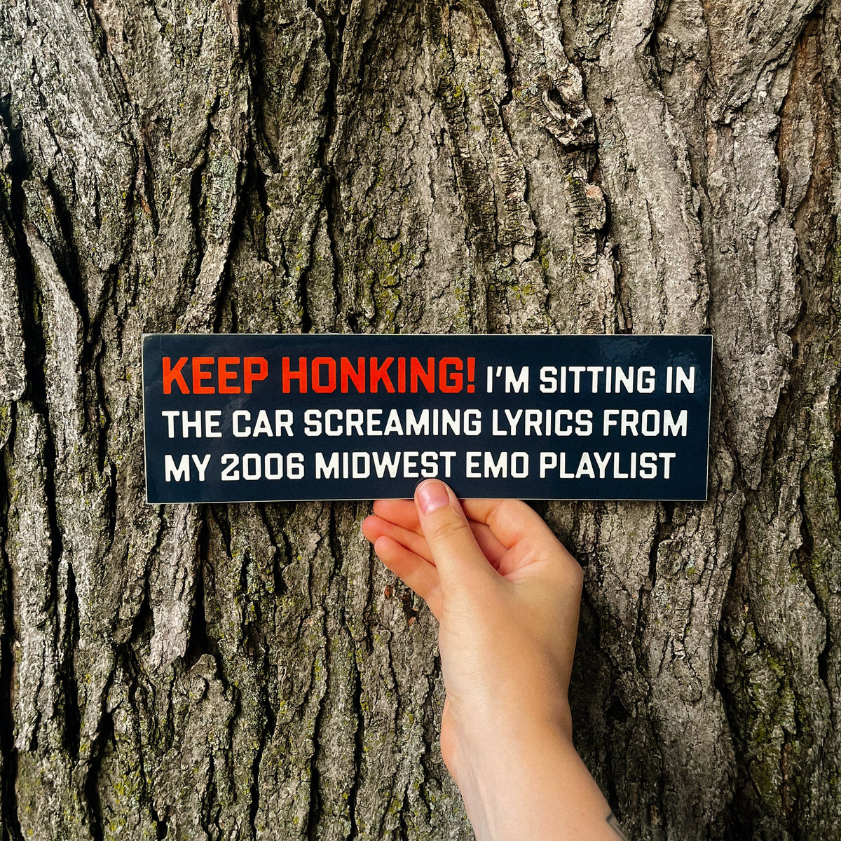 A hand holds a rectangular black novelty sign against a textured tree trunk. The sign has orange and white text that reads: 'KEEP HONKING! I'M SITTING IN THE CAR SCREAMING LYRICS FROM MY 2006 MIDWEST EMO PLAYLIST'.