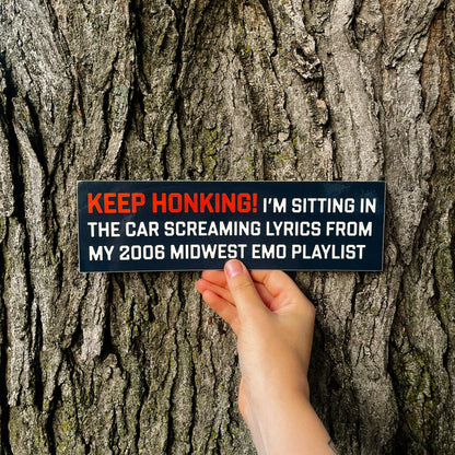A hand holds a rectangular black novelty sign against a textured tree trunk. The sign has orange and white text that reads: 'KEEP HONKING! I'M SITTING IN THE CAR SCREAMING LYRICS FROM MY 2006 MIDWEST EMO PLAYLIST'.