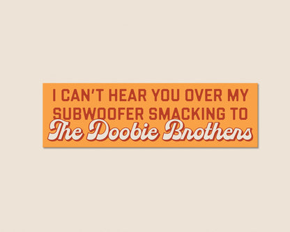 A rectangular orange sign with retro-style text reading 'I CAN'T HEAR YOU OVER MY SUBWOOFER SMACKING TO The Doobie Brothers' in red and white lettering, centered on a light beige background.